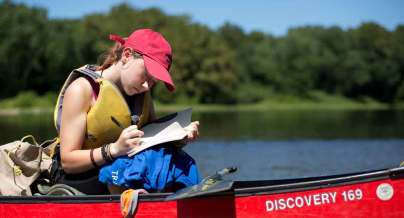 A person wearing a life jacket sits in a canoe writing something. There is a tree-lined shore in the background. 